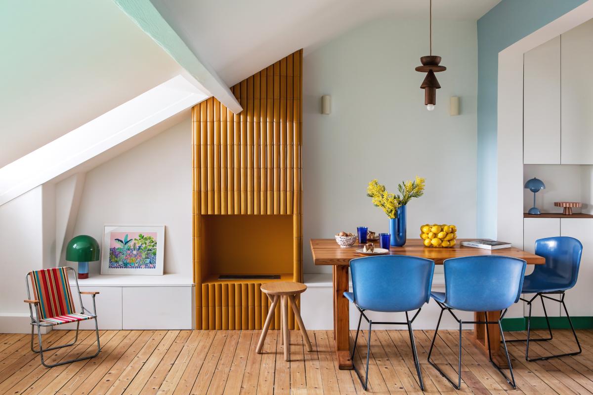 Wohnzimmer mit Kachelofen, Tisch und bunten Stühlen.