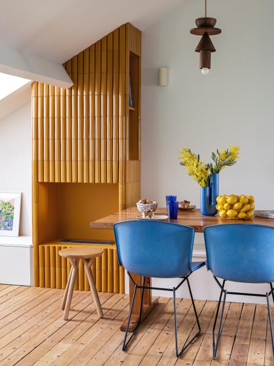 Wohnzimmer mit Kachelofen, Tisch und bunten Stühlen.